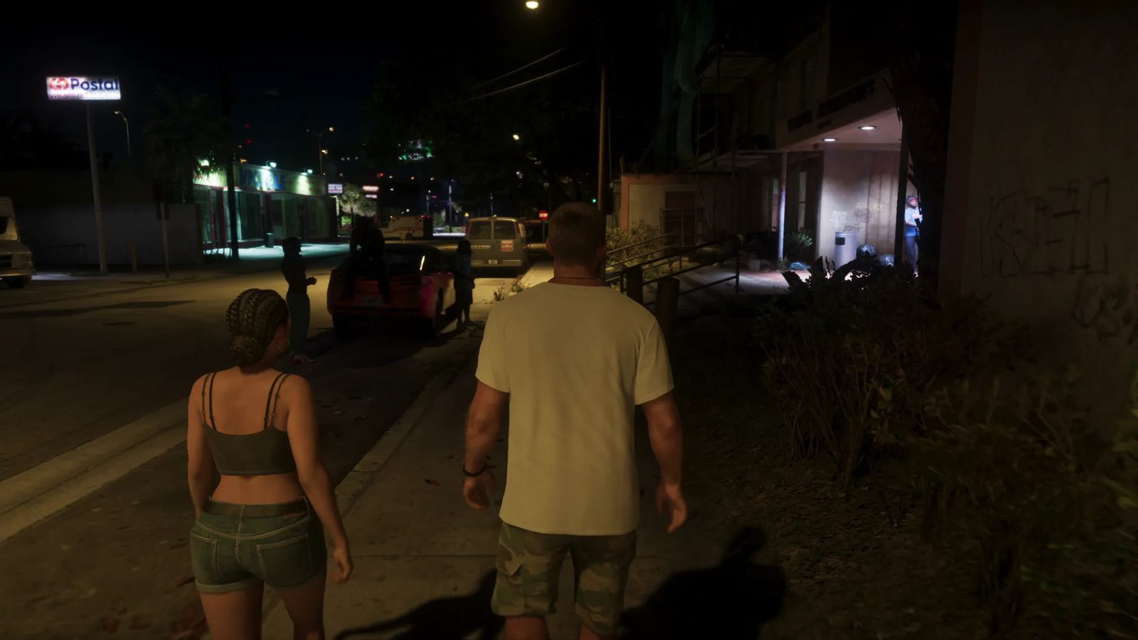 A woman with braids and a man in a white T-shirt walk along a dark city pavement at night, past a parked red car and a van.