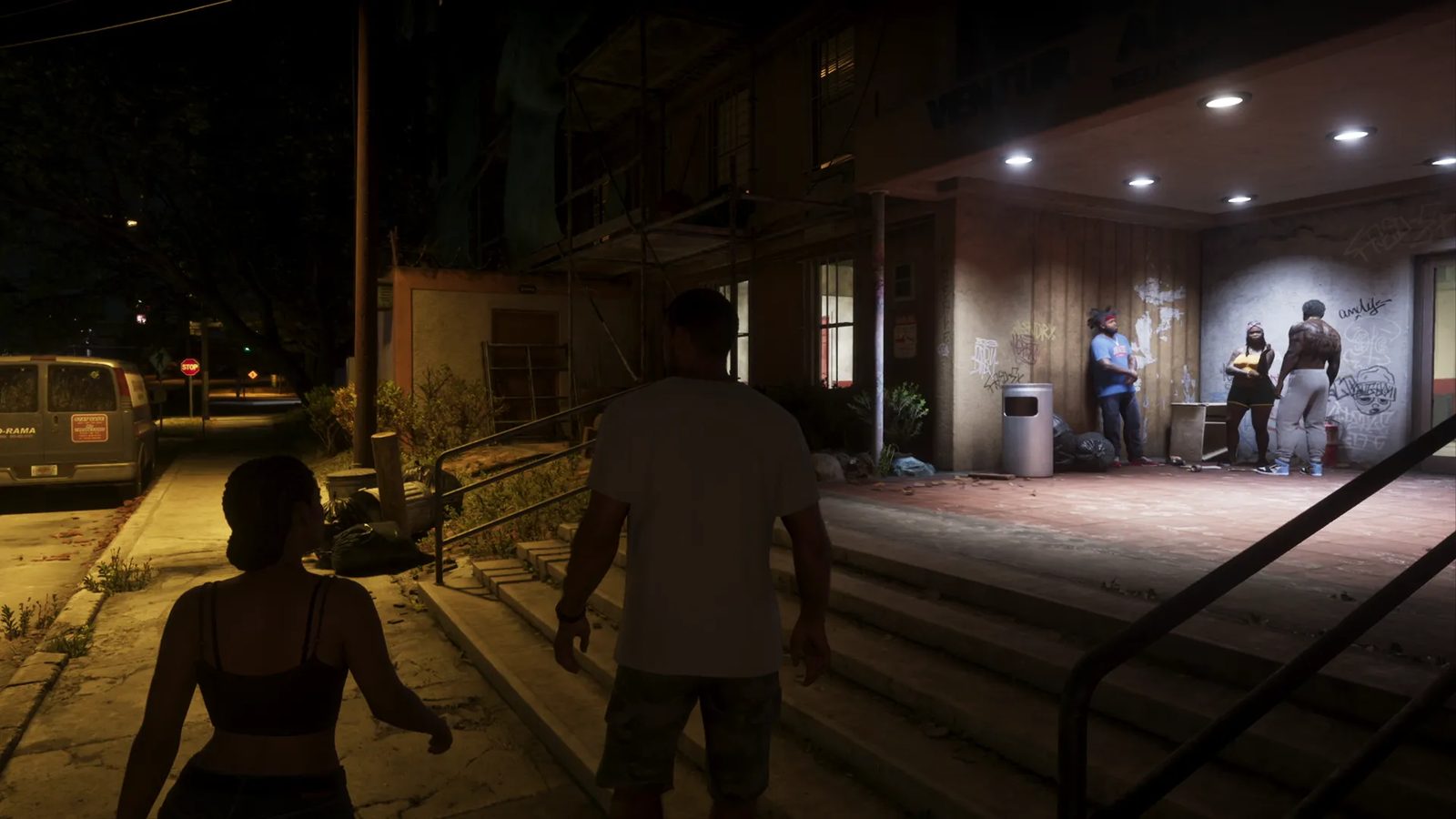 Jason Duval and Lucia Caminos walk along a night-time pavement towards a graffiti-covered building entrance where three people stand under lights.