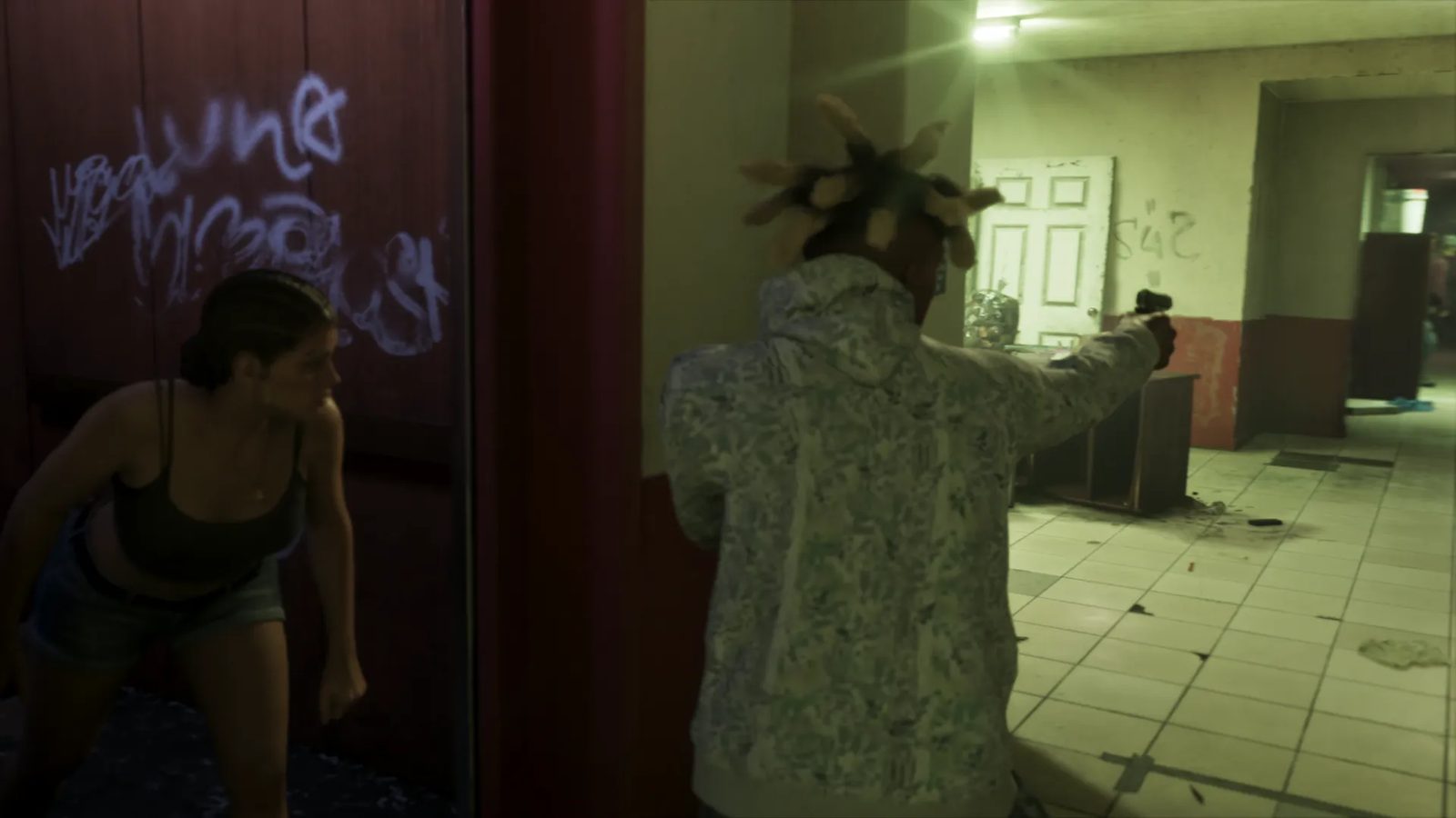 Lucia Caminos crouches beside a red doorway as a man in a patterned hoodie aims a handgun down a grubby, dimly lit tiled corridor.