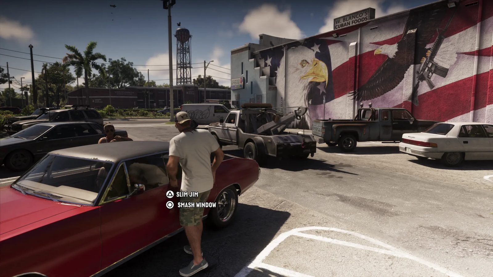 A man in a white T-shirt and camouflage shorts stands by a red car in a car park, beside a tow truck and a wall mural of an eagle and American flag.