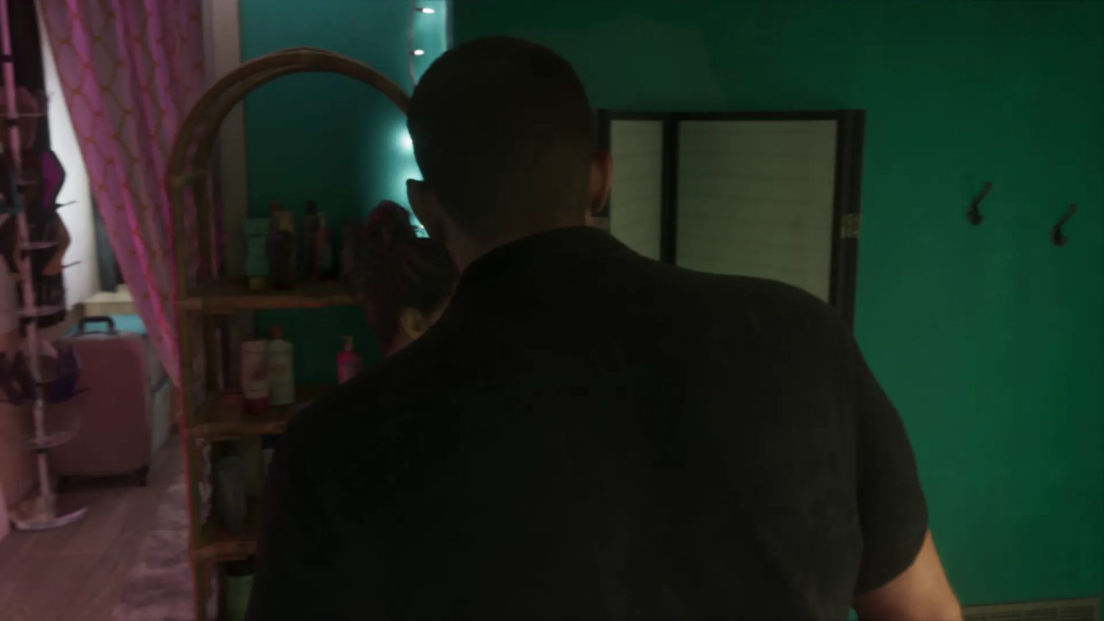 A broad-shouldered man in a dark shirt, seen from behind, stands in a dim teal bedroom beside a cane shelf, with a woman partly hidden beyond him.