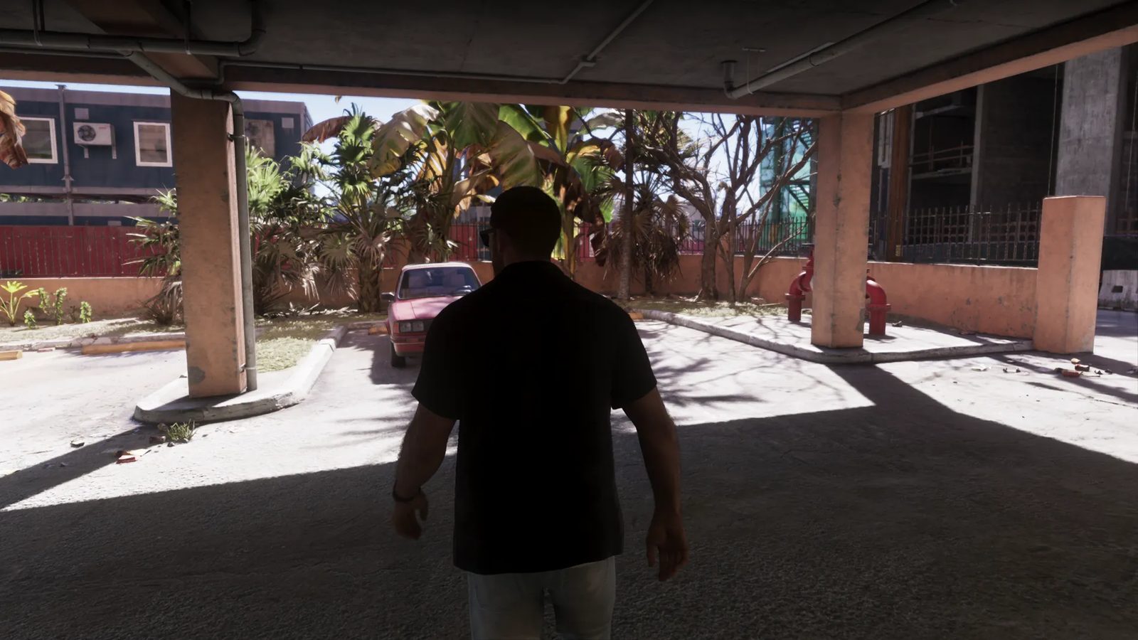 A man in a black shirt seen from behind walks through a shaded covered car park towards a pink car, with palm trees and a red wall beyond.