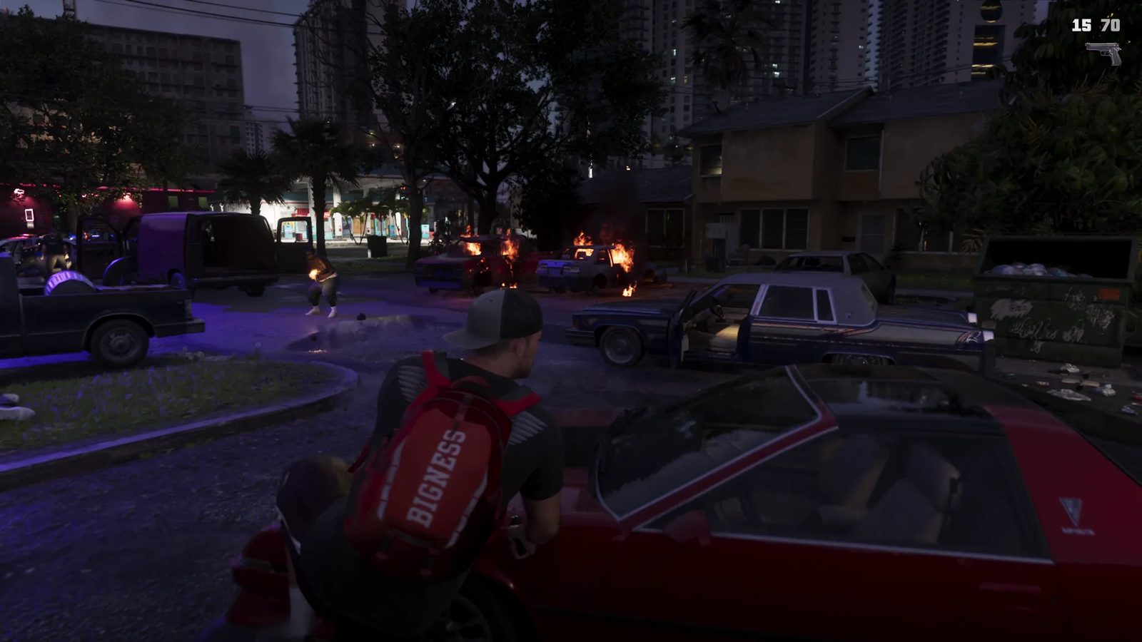 A man in a grey cap and red BIGNESS backpack crouches behind a red car on a night street where two cars burn and a man stands in flames nearby.