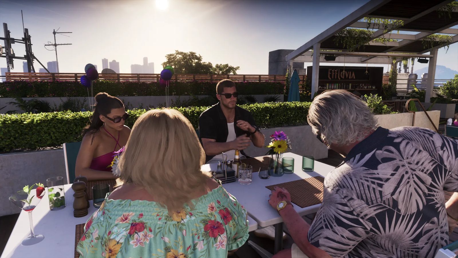 Four people sit at a white table on a sunny rooftop terrace, with a sign reading Effluvia behind them and a city skyline in the distance.