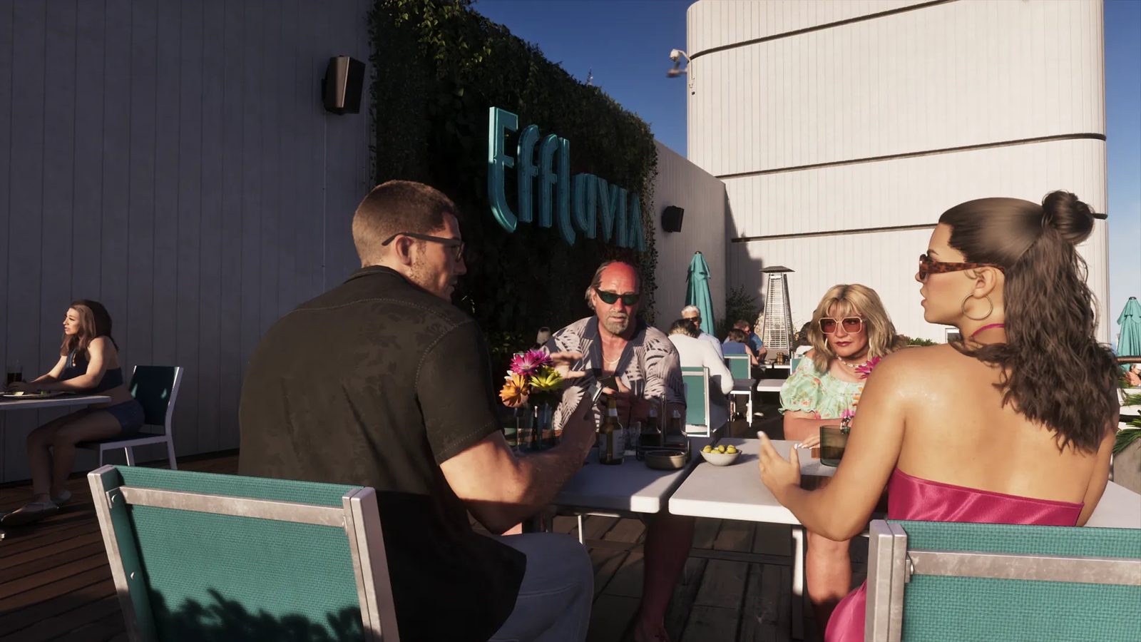 Four people talk around a white table on a sunny rooftop terrace beneath a teal Effluvia sign, with teal chairs and a woman seated alone behind them.