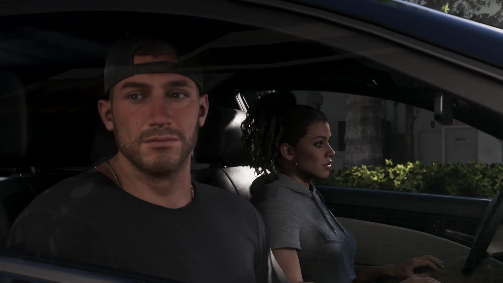 Jason Duval, in a backwards cap and dark t-shirt, sits in a car beside Lucia Caminos, who looks out of the window towards a hedge-lined street.