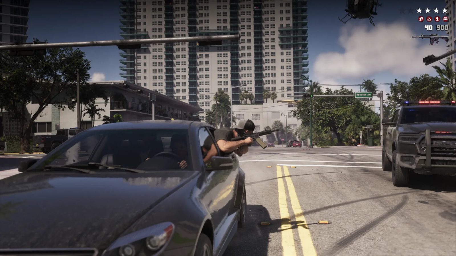 A man in a cap leans out of a grey car window firing a rifle at a police pickup truck on a sunny city street, with a helicopter overhead.