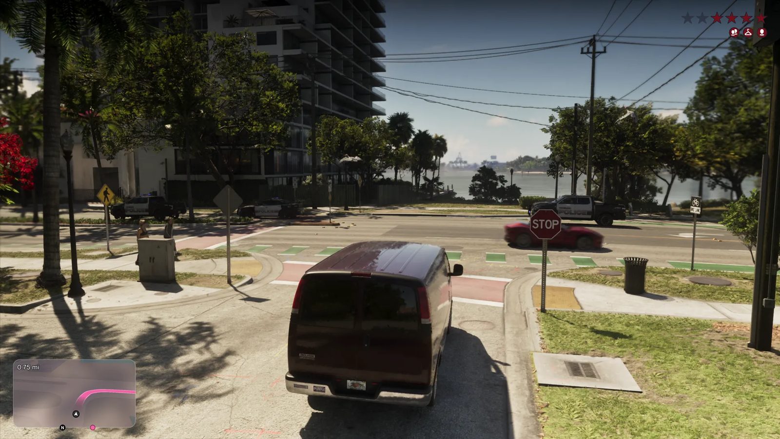 A dark red van on a sunlit street corner, with police vehicles and a red sports car ahead, a tower block on the left and a bay beyond, with a four-star wanted level.