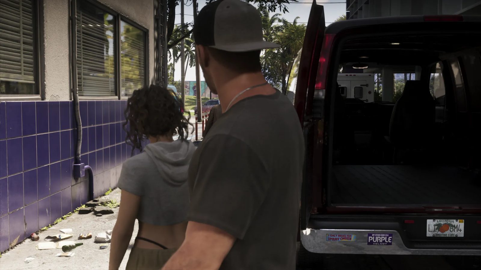 A man in a grey cap and a curly-haired woman walk past a purple-tiled wall towards a dark red van with its rear doors open on a sunny street.