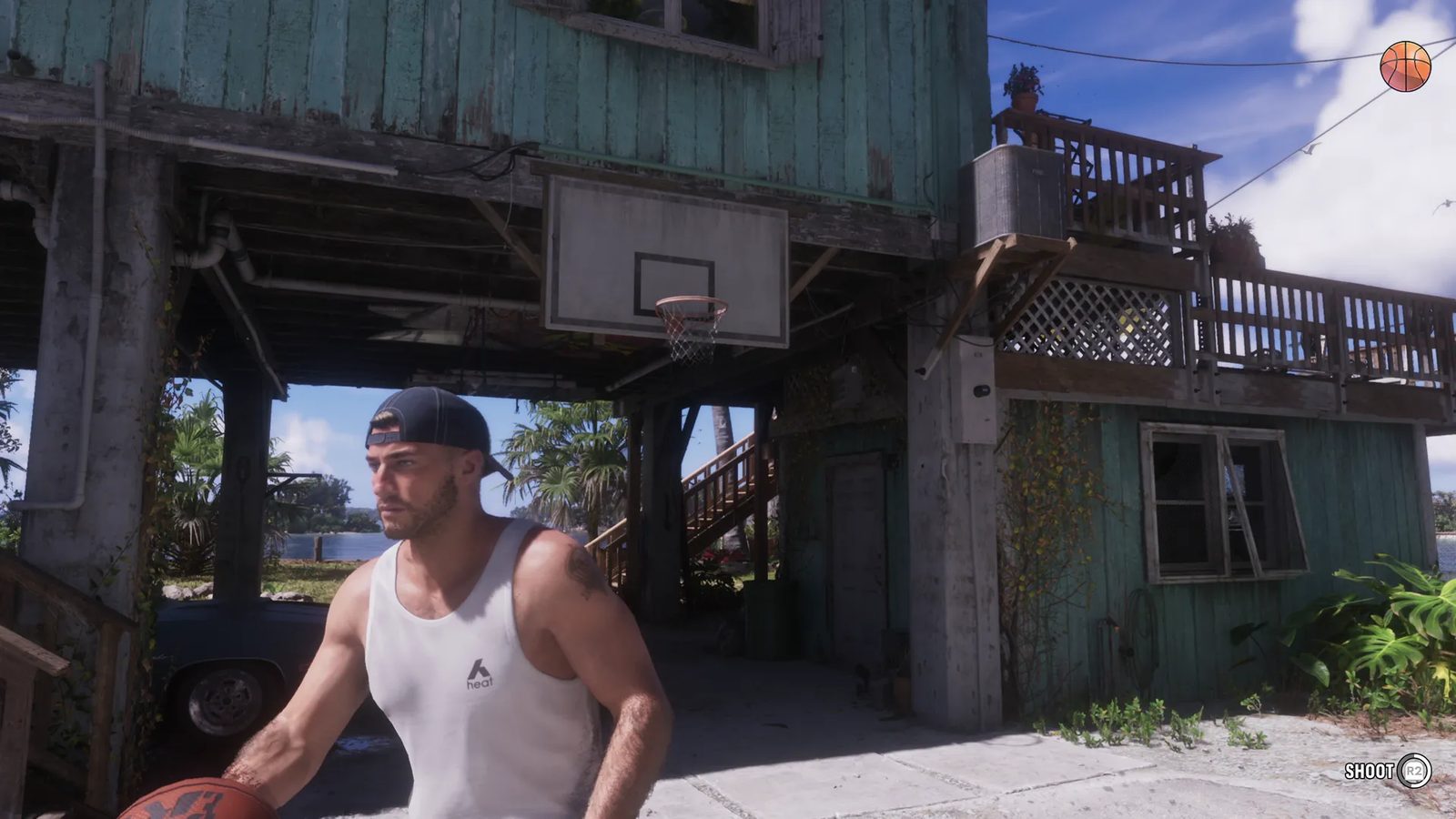 Jason Duval, in a white vest and backwards cap, holds a basketball beside a hoop under a weathered turquoise wooden house on stilts.
