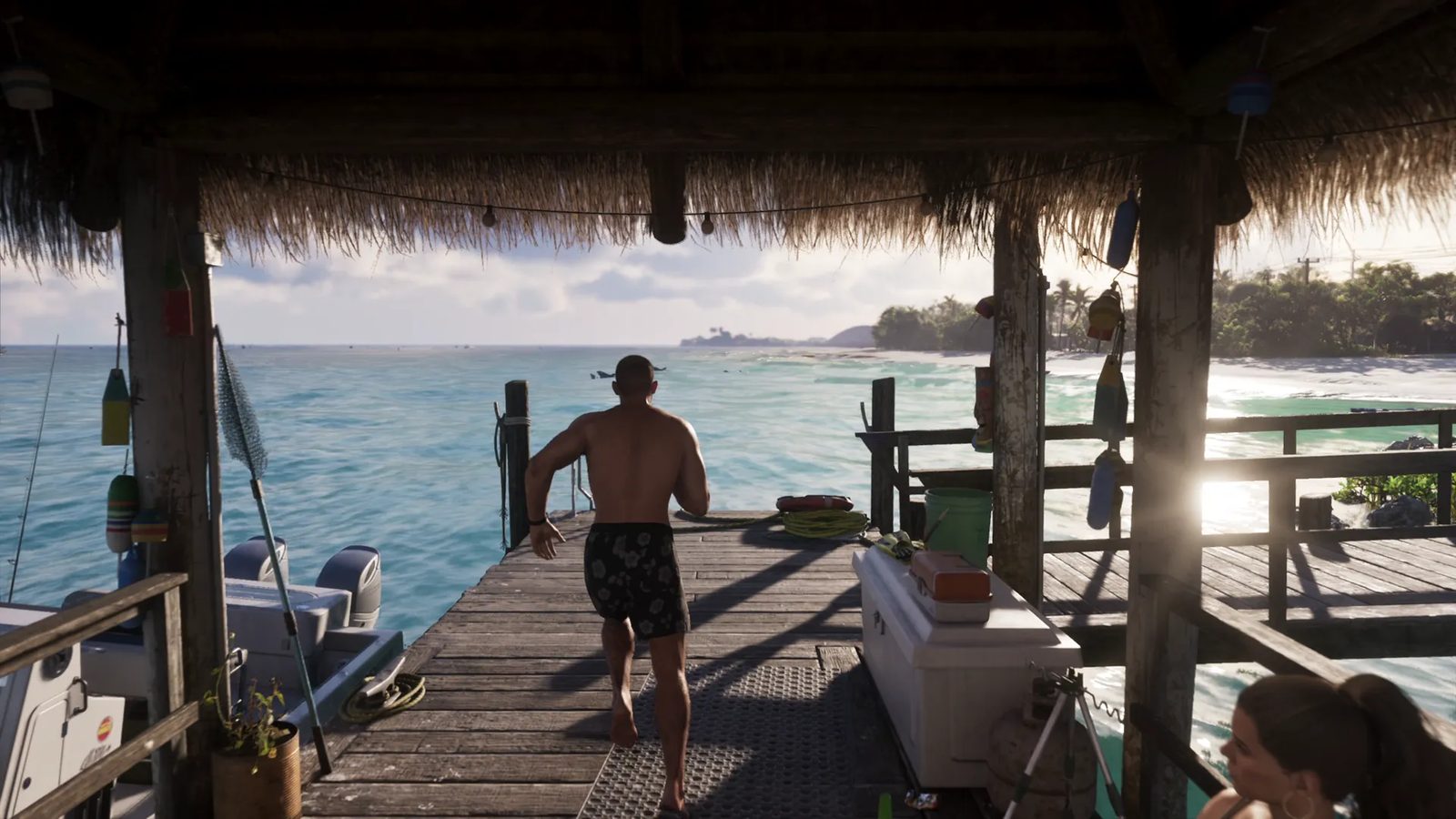 A shirtless man in floral swim shorts runs barefoot along a wooden jetty towards turquoise sea, beneath a thatched roof, in bright sunshine.