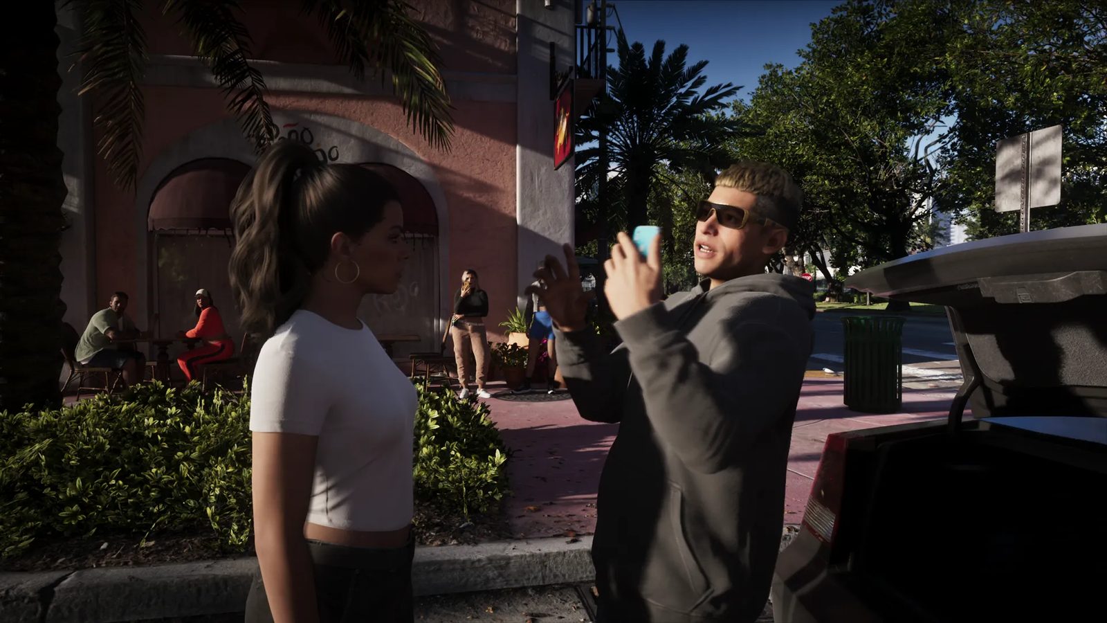 A man in sunglasses and a grey hoodie holds up a blue phone and gestures to a woman with a ponytail beside an open car boot on a sunny street.