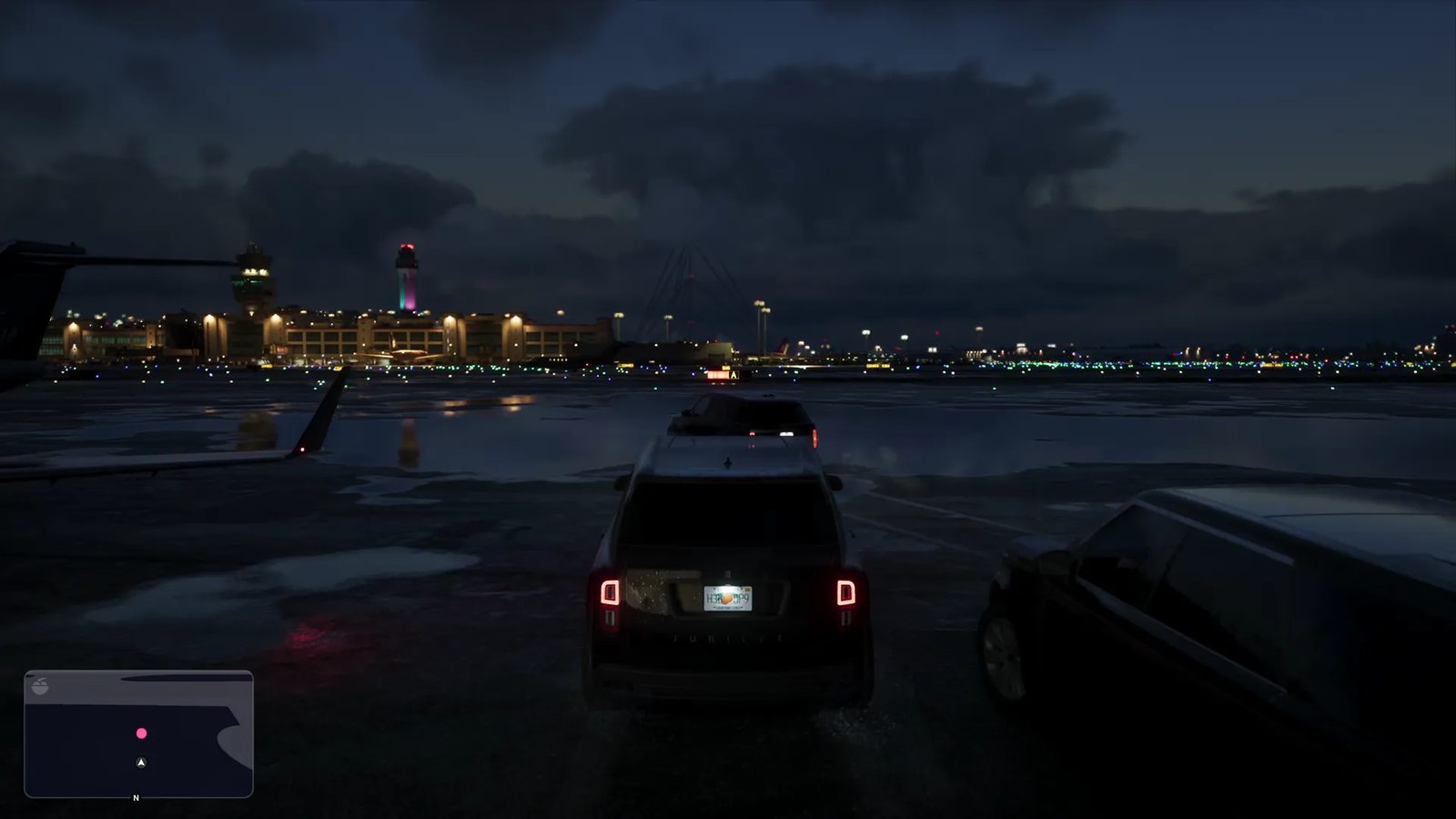 A dark SUV with lit tail lights is parked on a wet airport apron at night, facing distant terminal buildings and control towers under a cloudy sky.