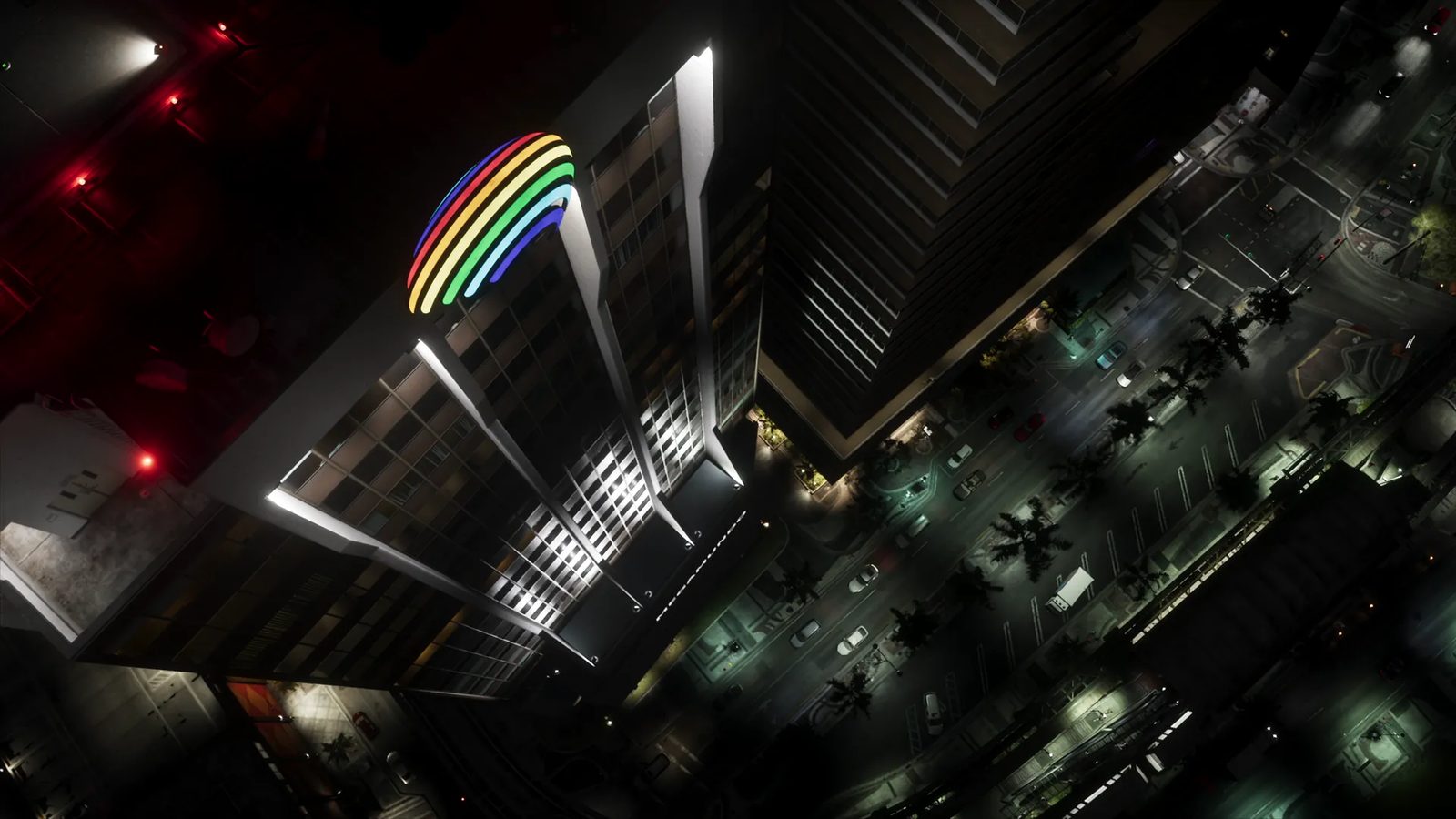 A high aerial view at night of a tall white-trimmed skyscraper with a rainbow-coloured neon sign on top, above a palm-lined city street with traffic.