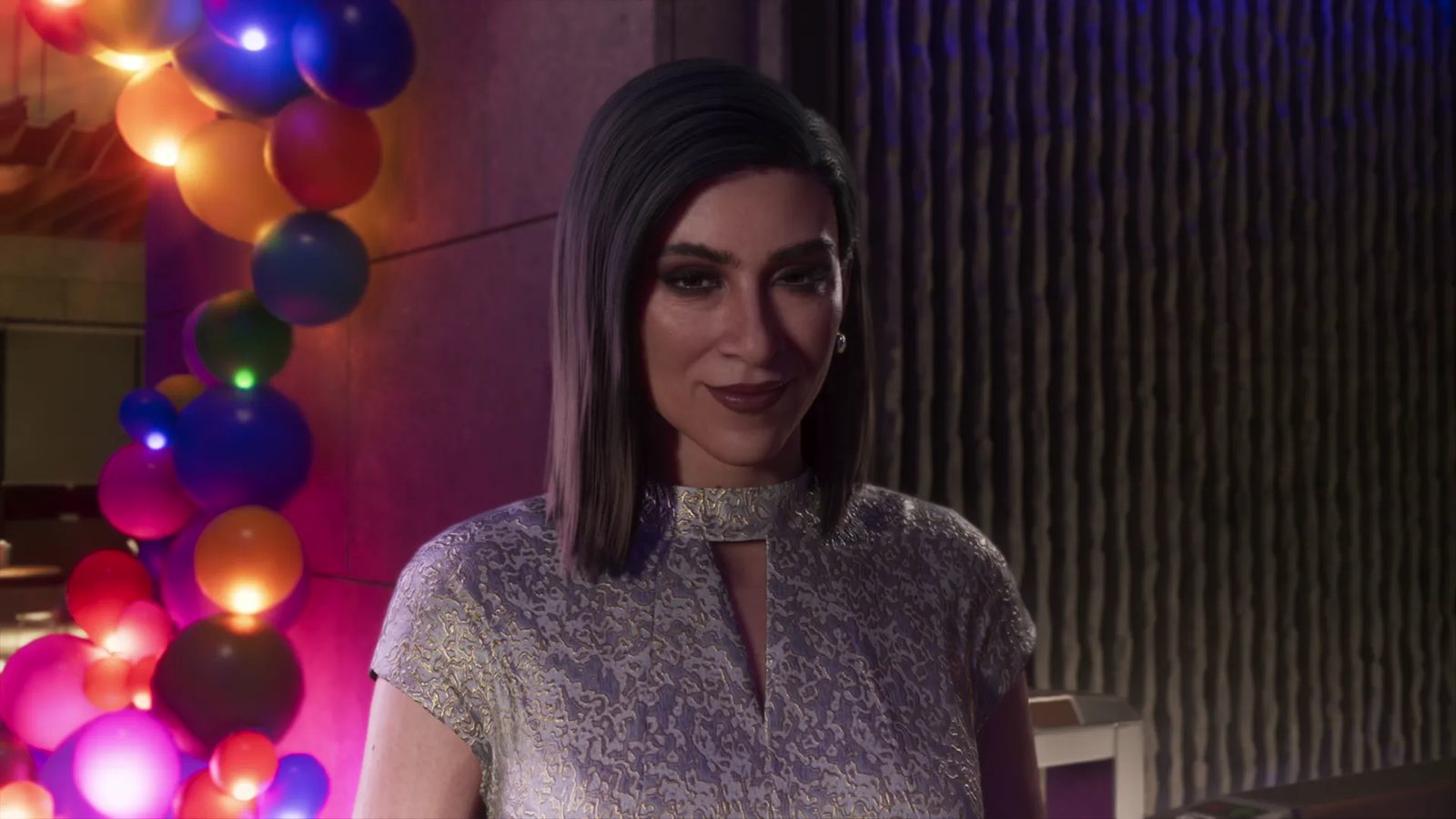 A smiling woman with a sleek dark bob and a silver-gold patterned top stands beside a column of colourful balloons in a dimly lit, purple-toned room.