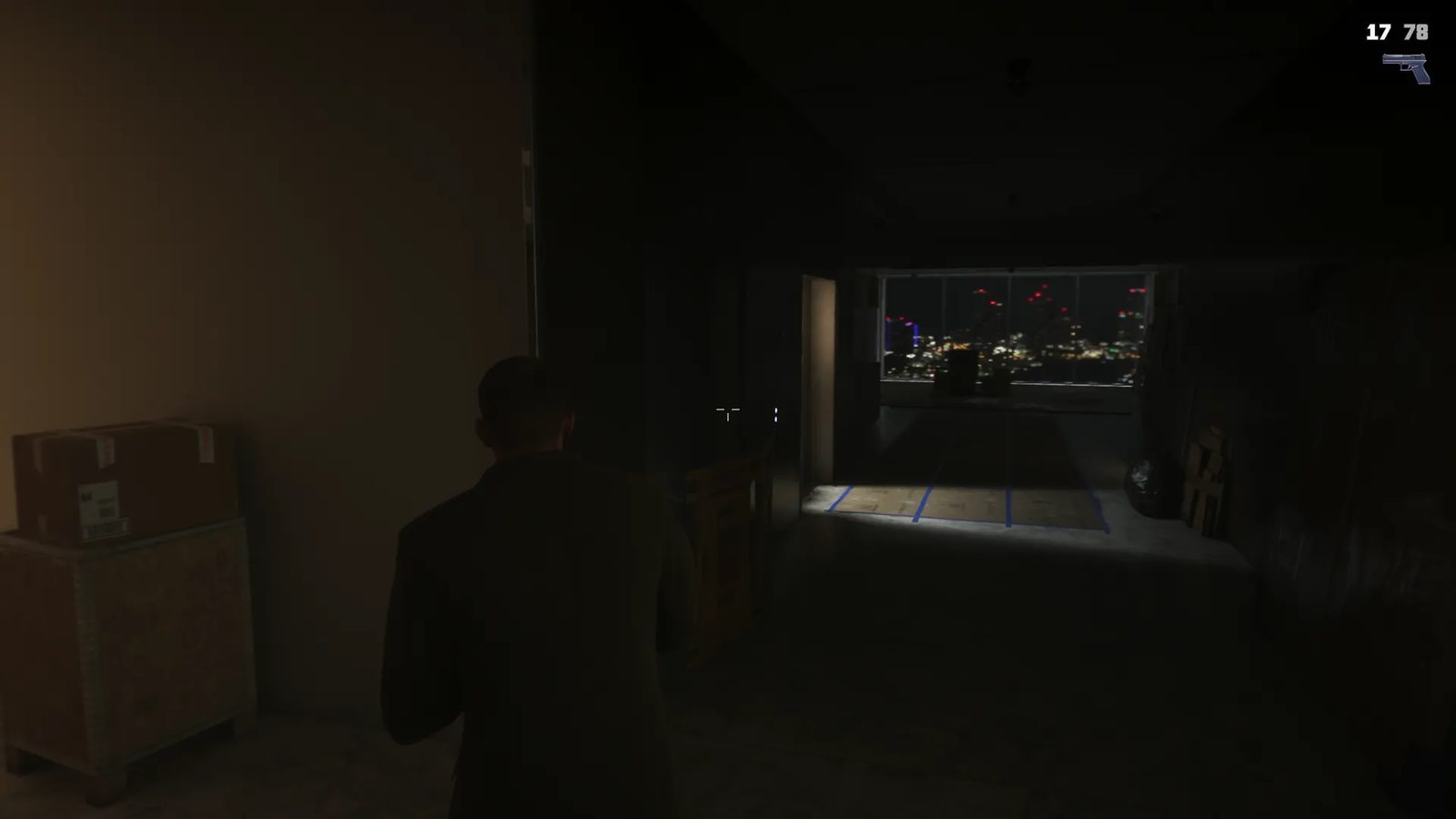 A man in a dark jacket, seen from behind, walks through a dim unfinished room towards a window overlooking a city lit up at night.