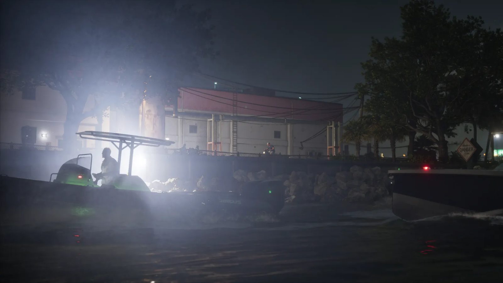 A silhouetted man steers a small open boat with a canopy at night, lit by a bright glare, beside a rocky shore and a warehouse.