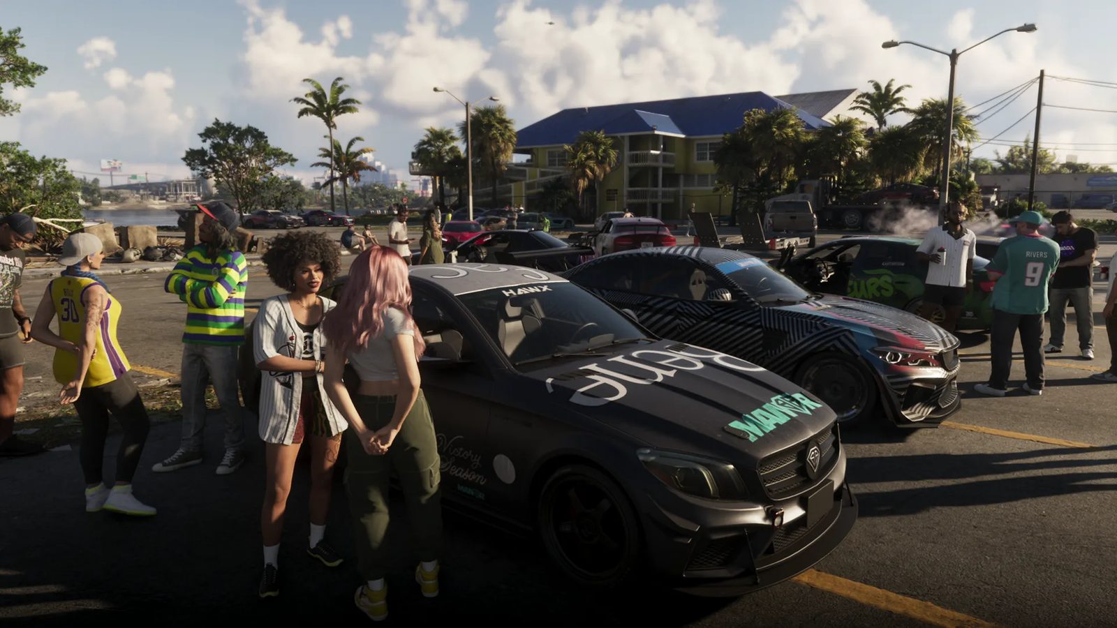 Groups of people stand around two modified black cars in a sunny waterside car park, with a yellow motel with a blue roof behind them.