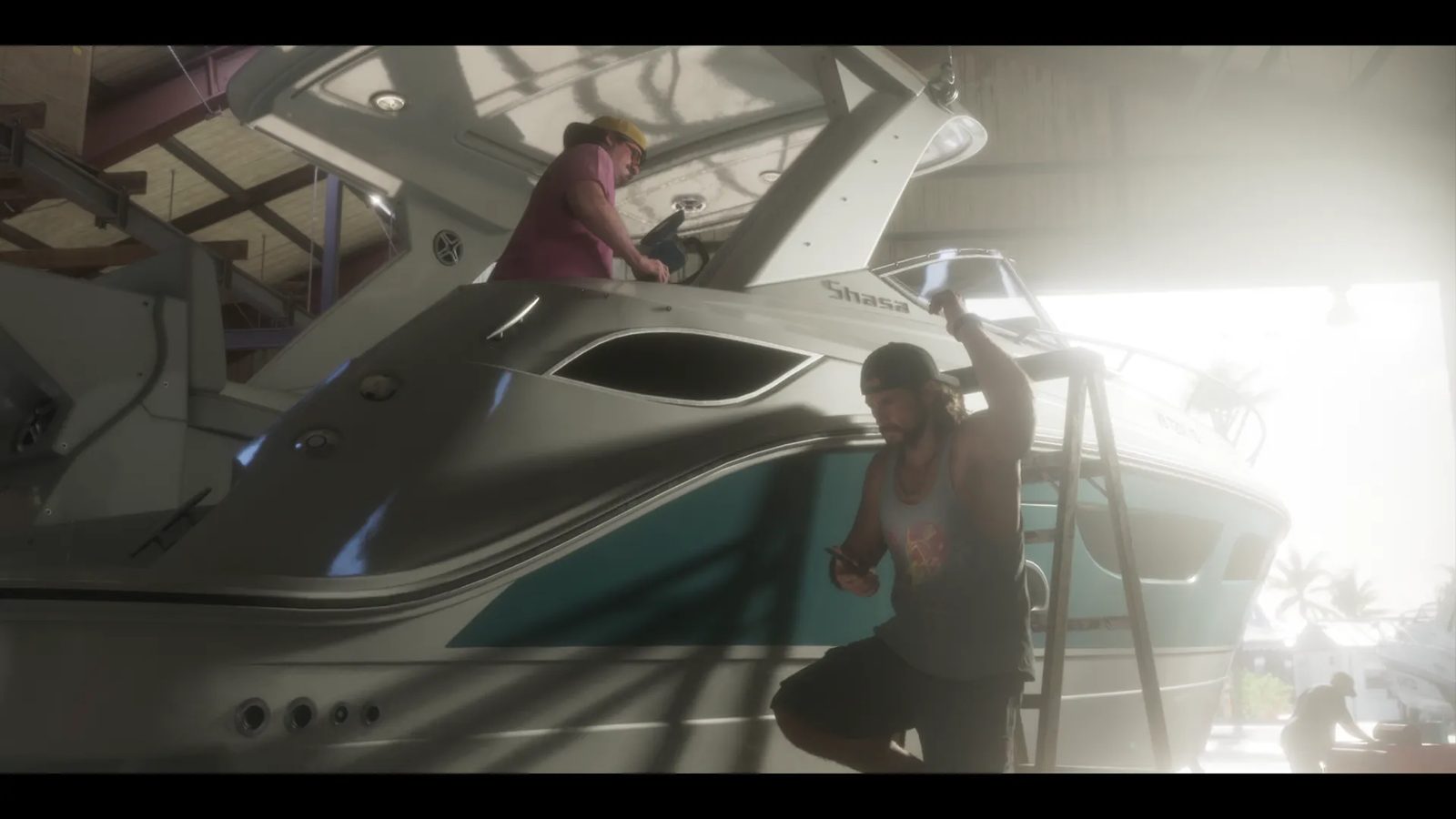 A man in a backwards cap checks his phone beside a ladder as another man in a yellow cap works on a white and teal boat in a boatyard shed.
