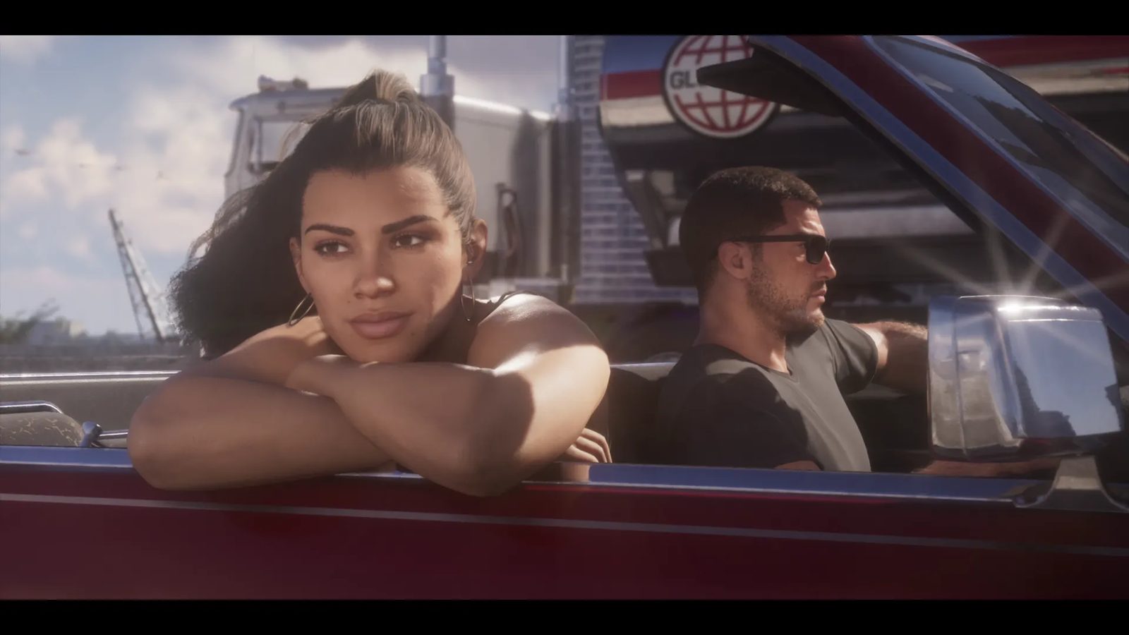 Lucia Caminos rests her arms on the door of a dark red convertible while Jason Duval, in sunglasses, drives beside a lorry on a sunny day.