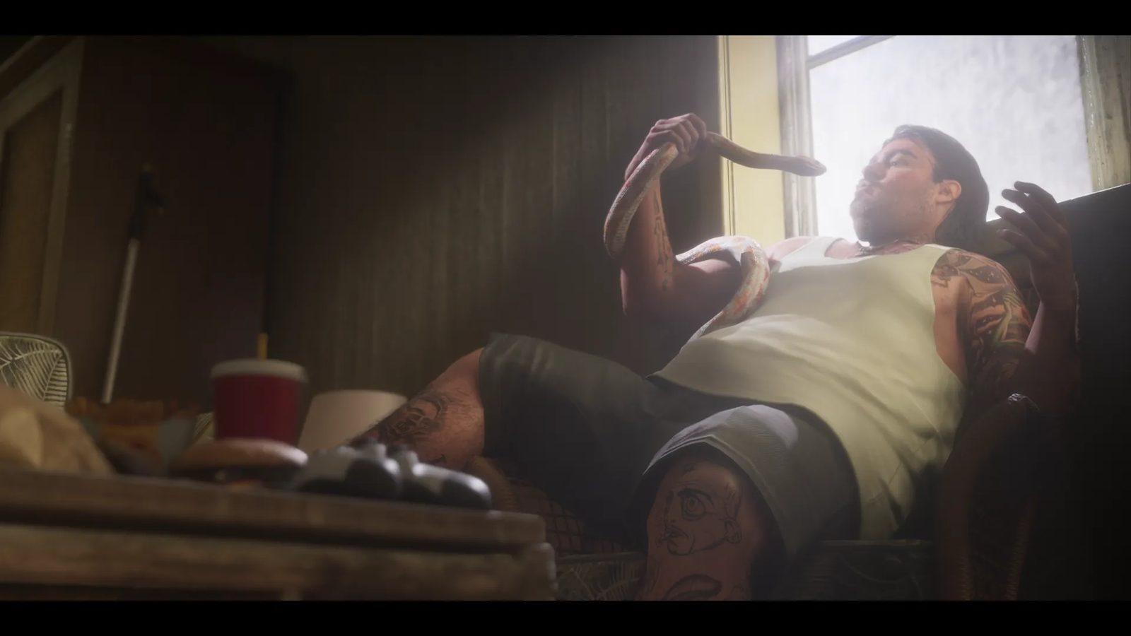 A heavily tattooed man in a white vest and grey shorts reclines in a chair by a bright window, holding a pale snake draped over his shoulder.