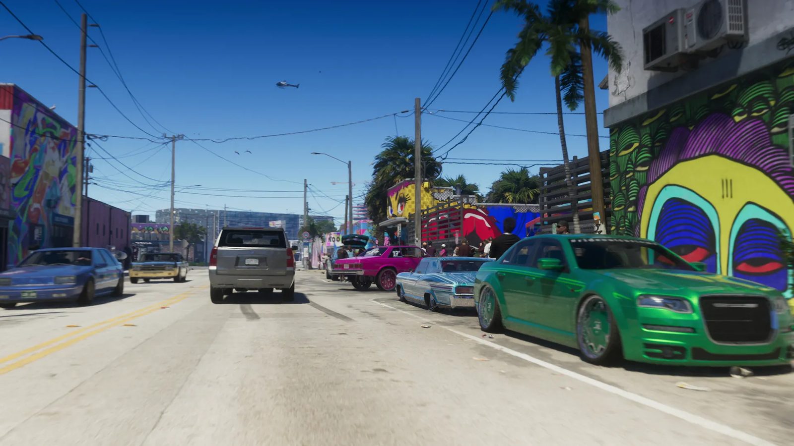 A lowered green saloon car and other custom cars park beside a mural-covered wall on a sunny street lined with palm trees, with a helicopter in the sky.