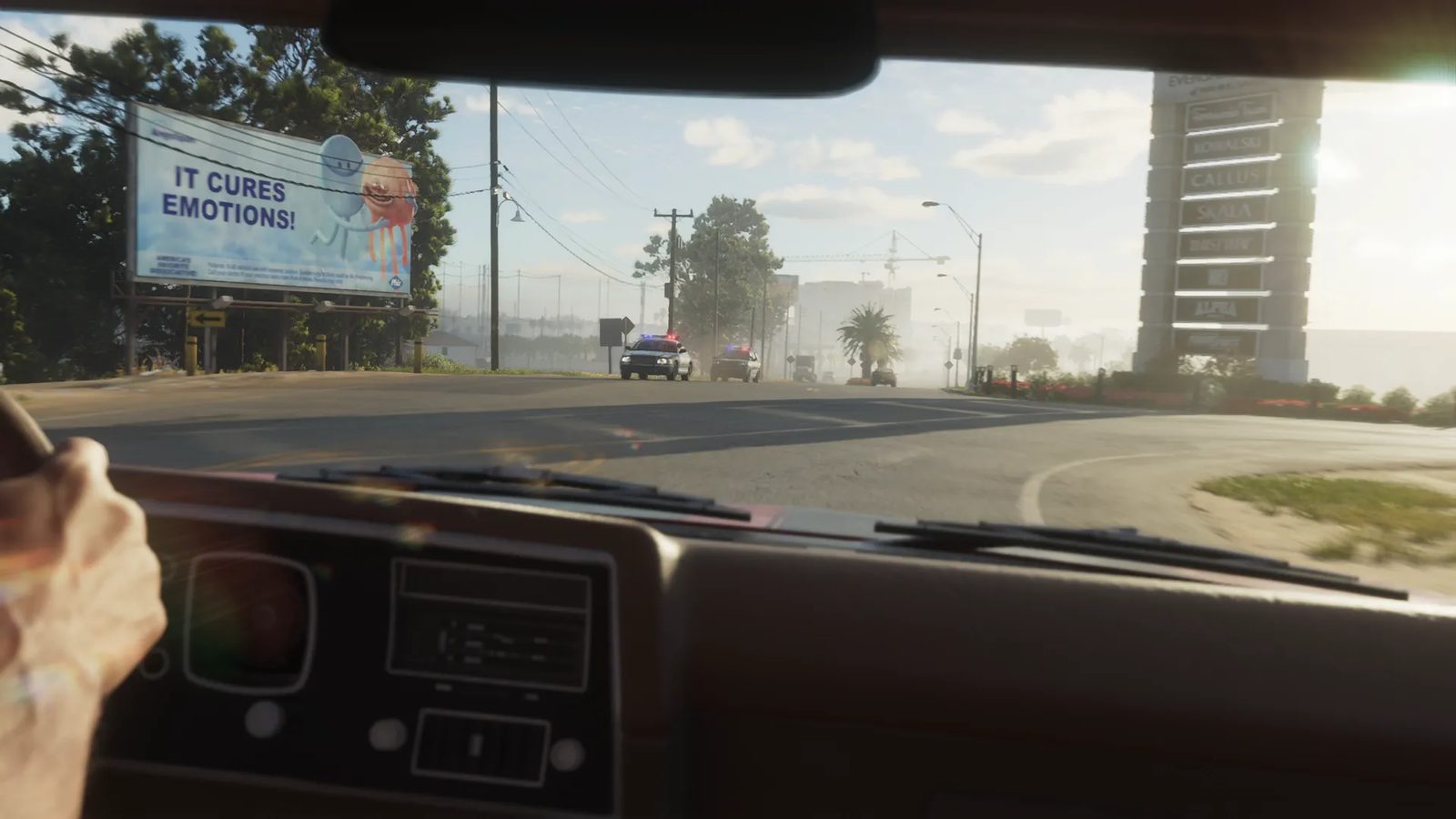 View from inside a car, one hand on the wheel, as two police cars with flashing lights approach along a sunlit road past a billboard reading "It cures emotions!"