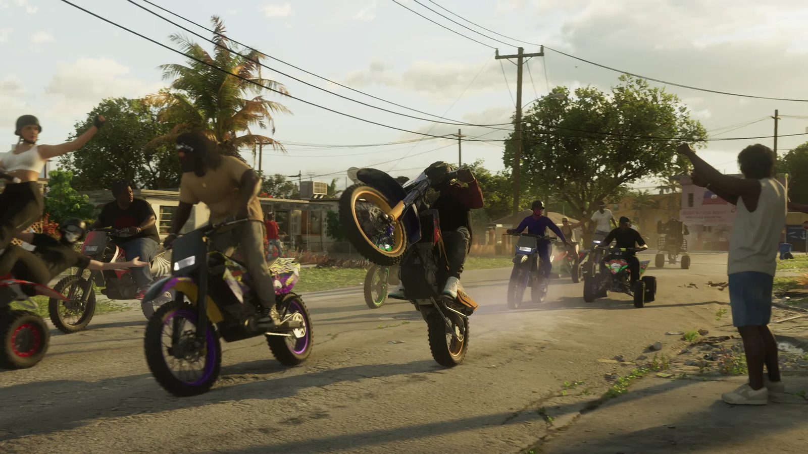 A rider pops a wheelie on a dirt bike on a sunlit residential street as other bikers and quad bikes follow and onlookers cheer.