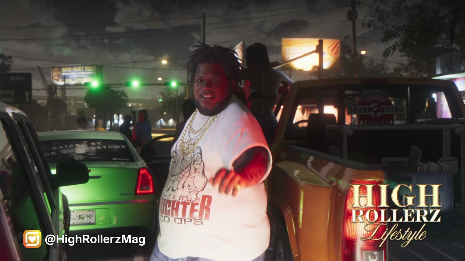 A man in a white T-shirt and gold chains gestures beside a green Schyster PMP 700 and an orange pickup at night, under green traffic lights.