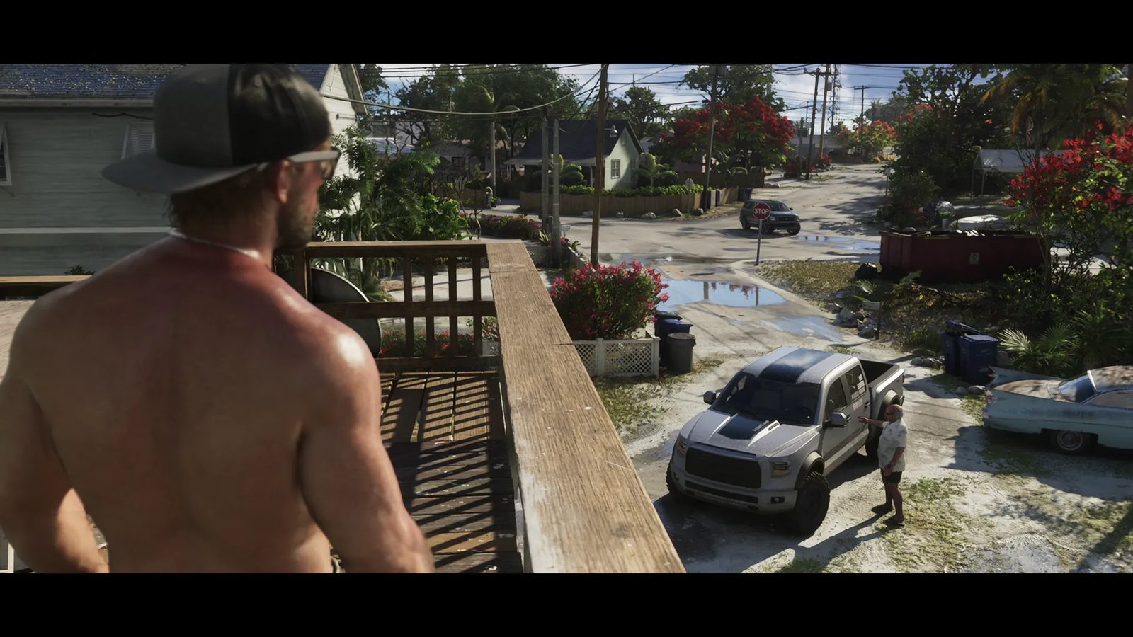 A shirtless man in a cap and sunglasses looks down from a wooden balcony at a silver pickup truck and a man in a white shirt on a sunny residential street.