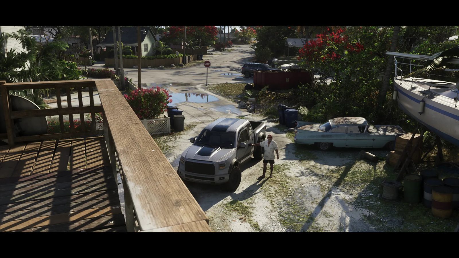 A man in a white floral shirt stands beside a silver pickup truck in a sandy yard, seen from a wooden balcony, with an old car and a boat nearby.