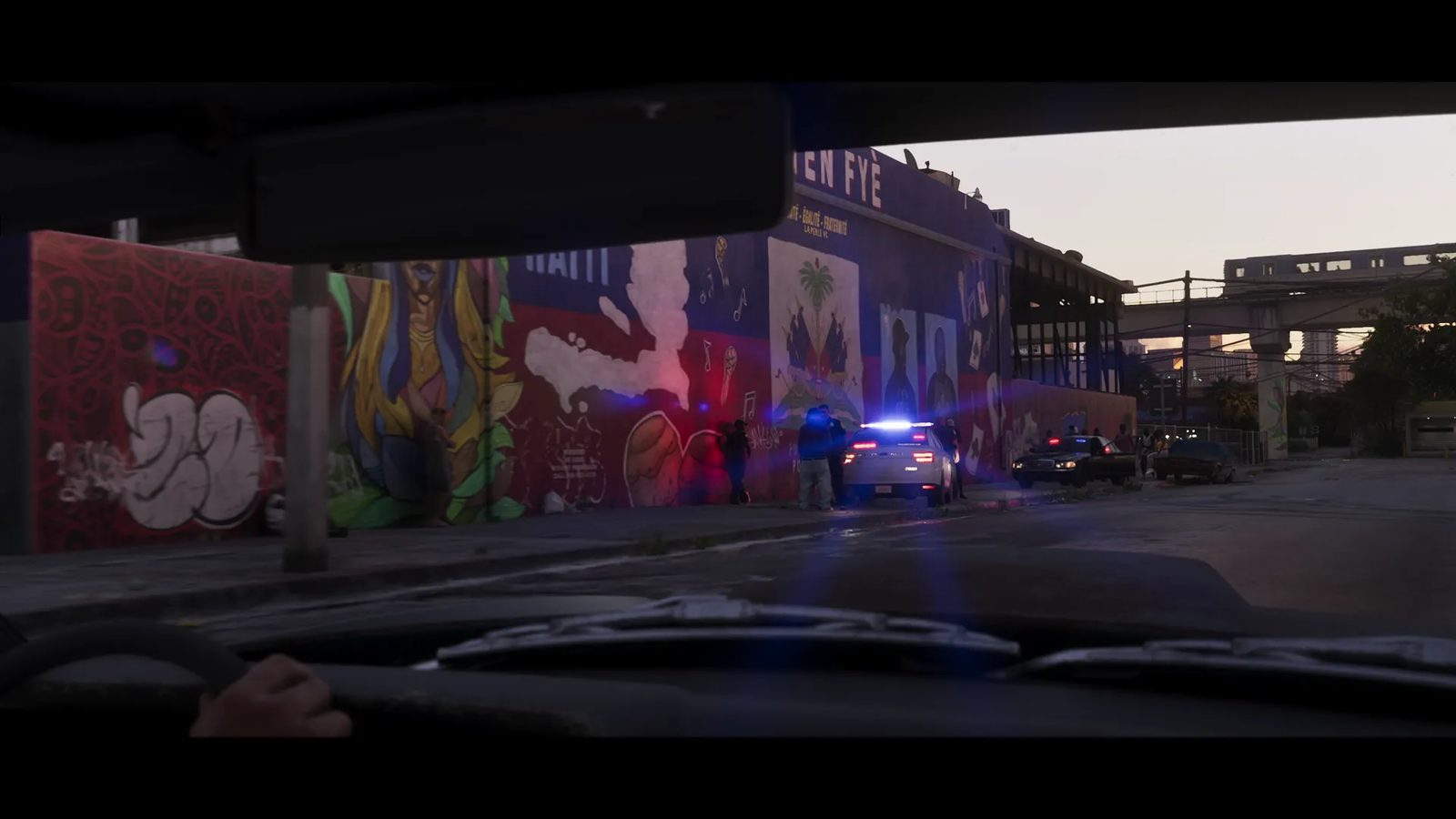 View from inside a car at dusk of a police car with flashing lights and officers by a large Haiti mural on a wall in La Perle.
