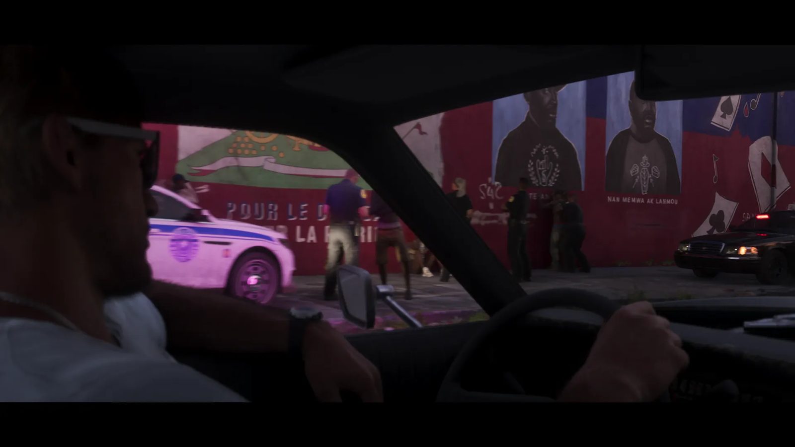 A man in sunglasses drives past police officers detaining people in front of a red mural at dusk, with a white police car and a dark police car nearby.