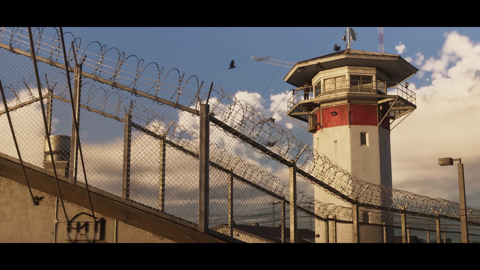 A red and white guard tower stands behind chain-link fences topped with razor wire, with a person on its balcony and a jet in the sky.