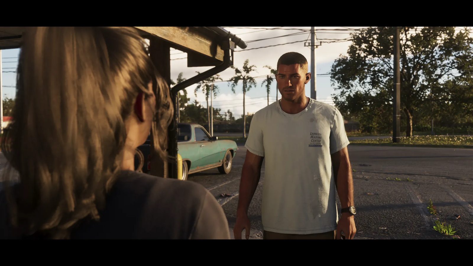 Jason Duval, in a pale grey t-shirt, stands in a car park facing a woman seen from behind, with a faded green car parked behind them.