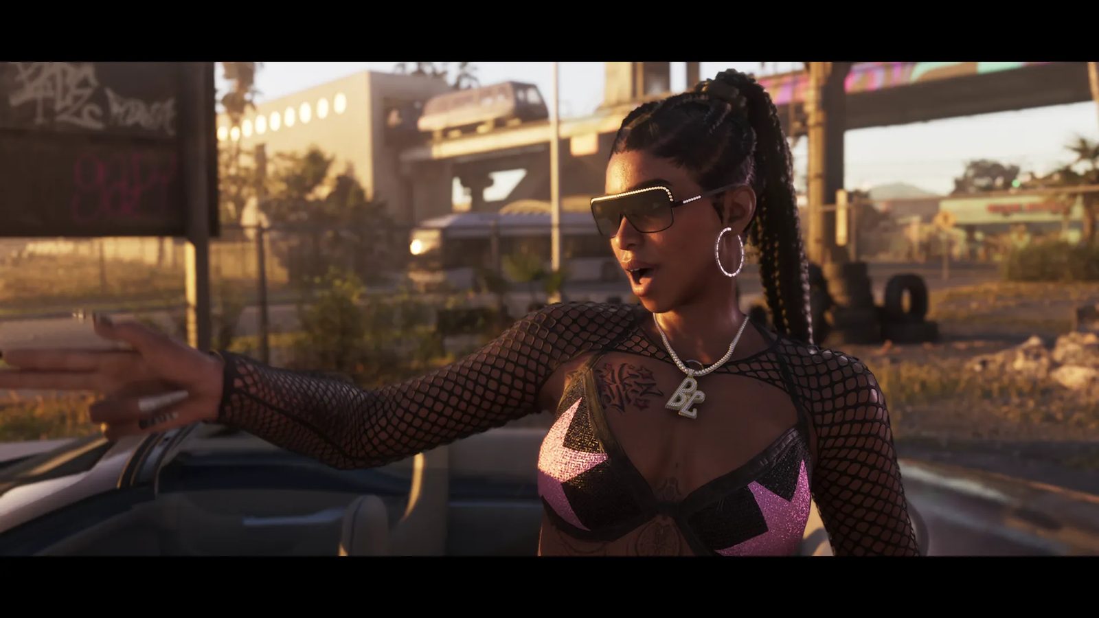 A woman in sunglasses, a fishnet top and a pink and black bra points out of an open-top car in warm evening light, with a raised train line behind.