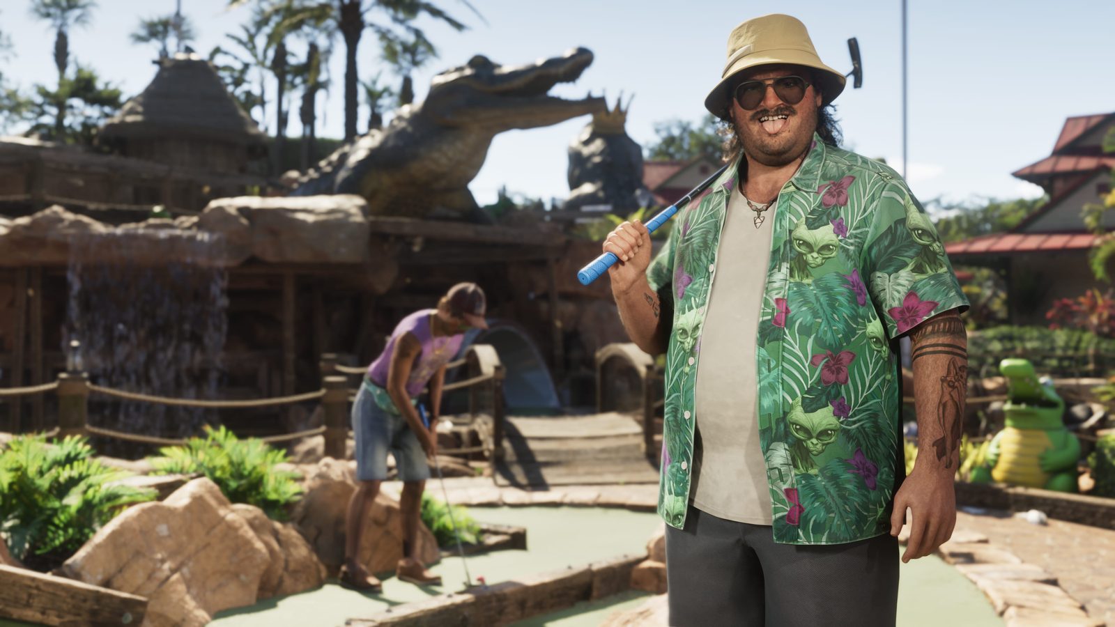 Cal Hampton, grinning in a bucket hat and alien-print shirt, rests a putter on his shoulder at a sunny crazy golf course with a giant crocodile statue.