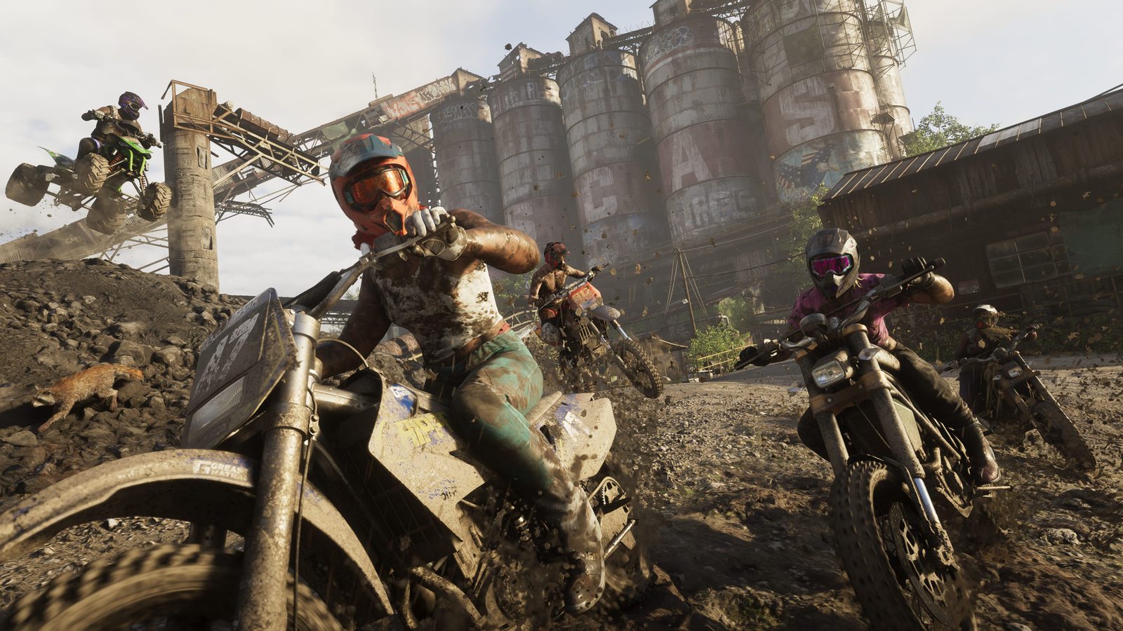 Muddy riders on dirt bikes race across a rubble-strewn track in front of tall, graffiti-covered concrete silos, with a quad bike airborne behind.