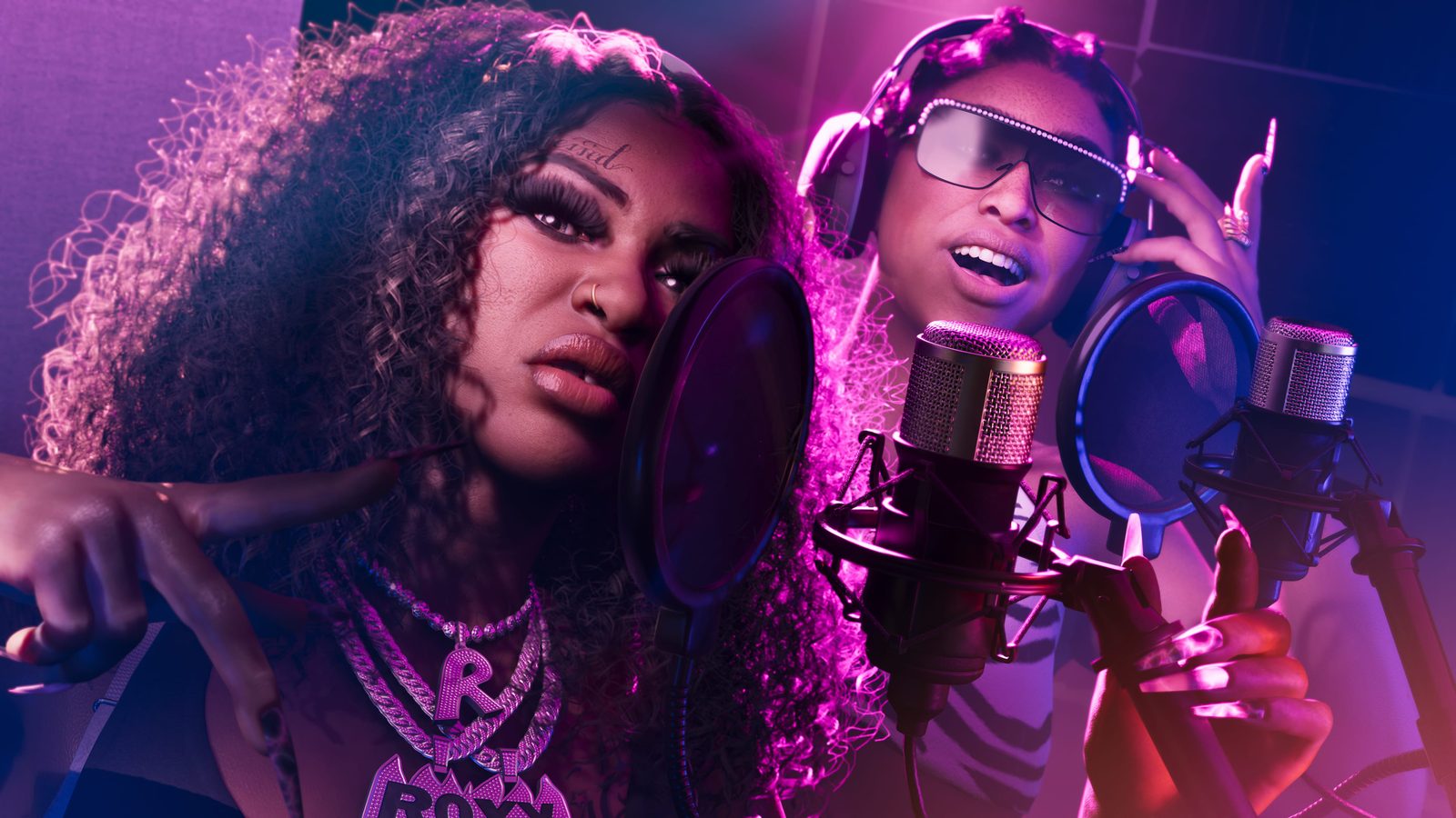 Two women in a recording studio rap into pop-filtered microphones, one wearing chunky chains, the other headphones and jewelled sunglasses, in purple light.
