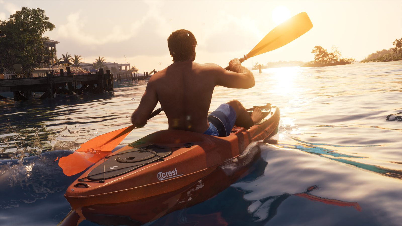 A shirtless man paddles an orange Crest Kayak across calm water towards a wooden jetty and waterfront houses at sunset.