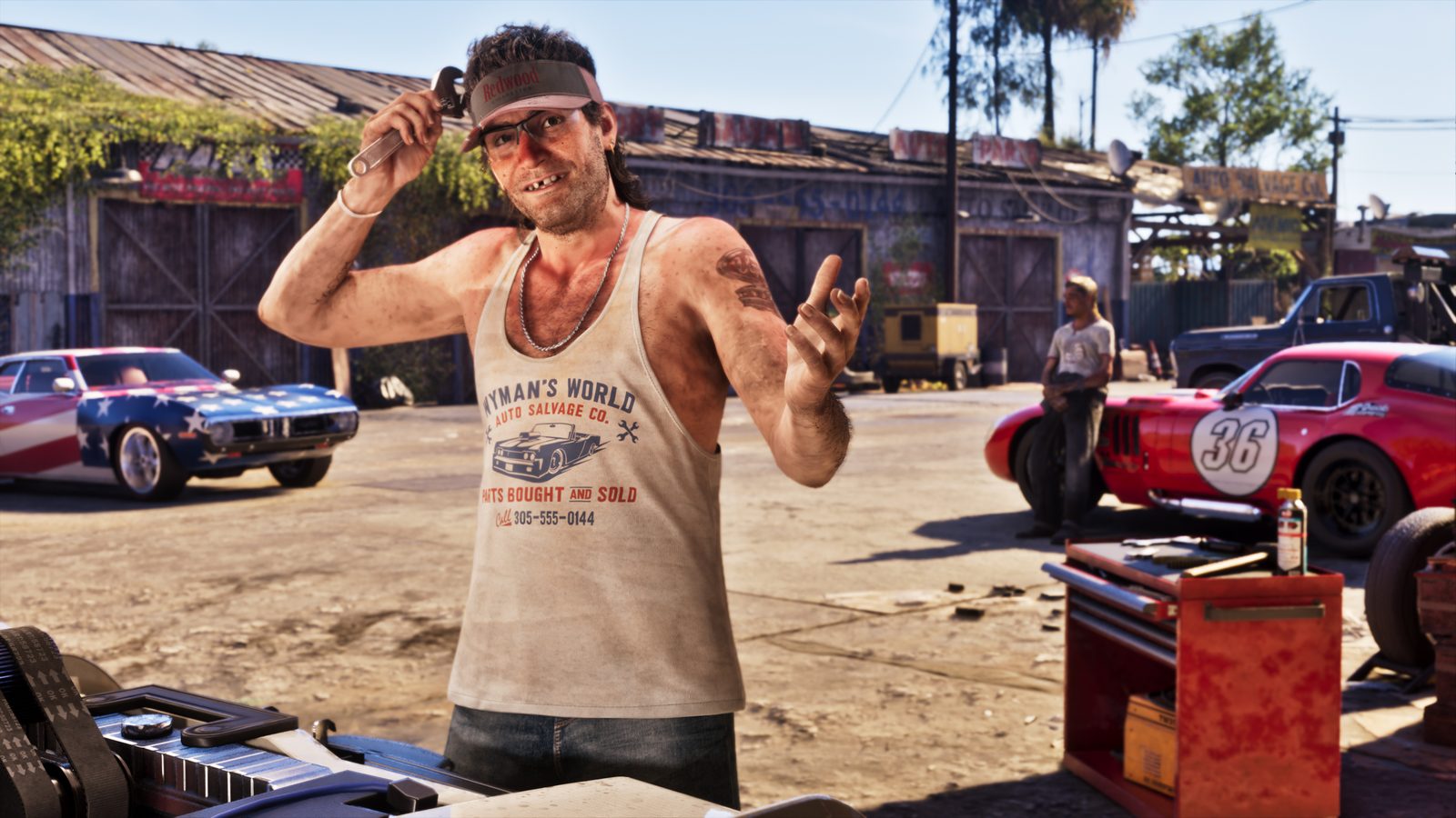 Wyman, a grinning man in a Redwood visor and vest, holds a wrench to his head beside classic cars in a sunny salvage yard.