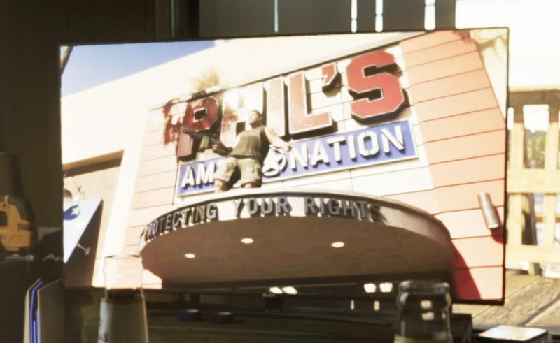 A man in a vest and cargo shorts stands on the awning of Phil's Ammu-Nation, holding a rifle, shown on a television screen in a room.
