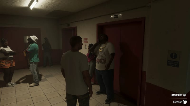 A man in a white t-shirt faces a group waiting in a dim tiled hallway by red lift doors, while a large man in gold chains talks on a phone.