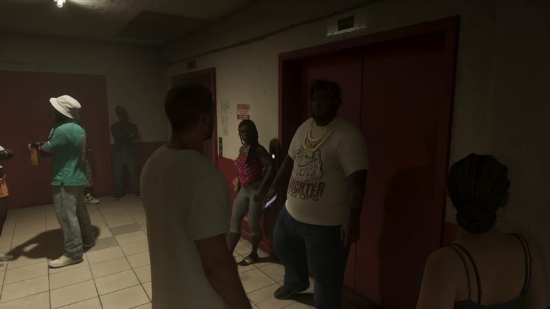 A group of people stand talking in a dim tiled lobby beside red lift doors, including a large man in a white T-shirt and gold chains.