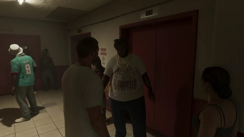 A group of people stand in a dim tiled lift lobby beside red lift doors, with a large man in a white slogan T-shirt and gold chains at the centre.