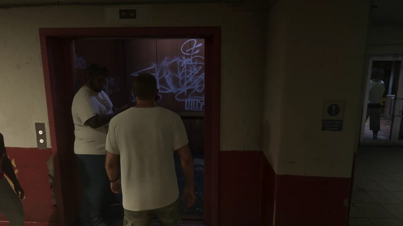 Two men in white T-shirts stand at an open lift with graffiti on its doors, in a dim hallway with red and cream walls.
