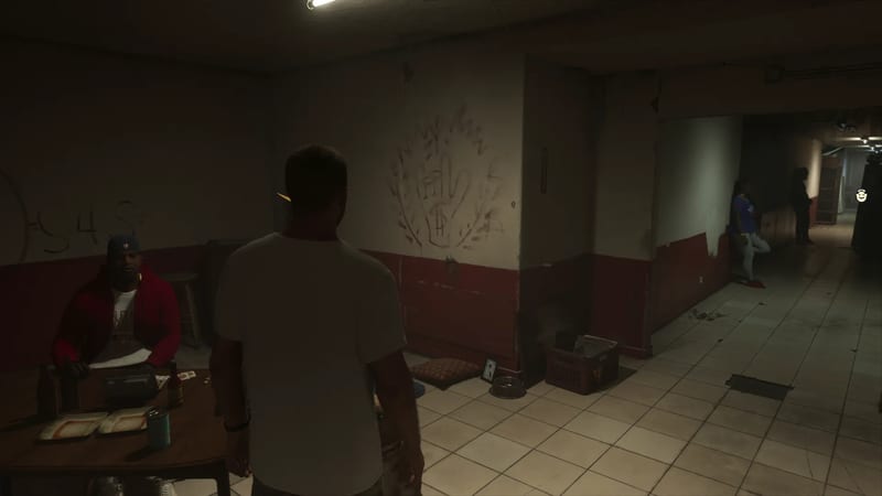 A man in a white T-shirt, seen from behind, stands in a dim tiled basement corridor facing a seated man in a red hoodie at a wooden table.