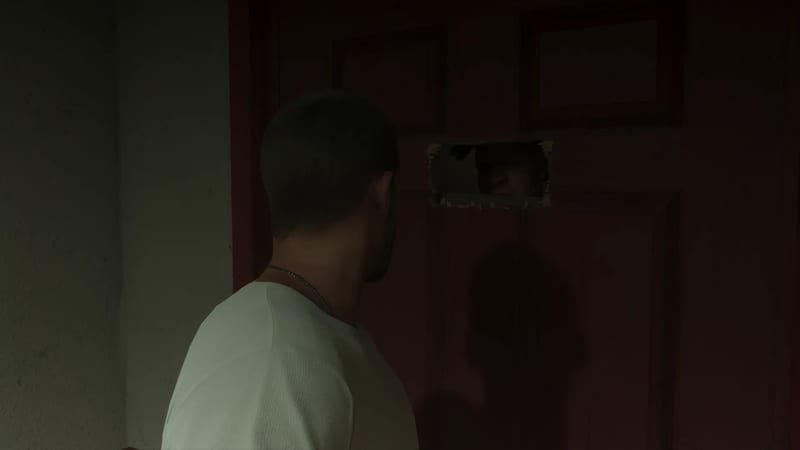 A short-haired man in a white top and chain necklace stands in a dim hallway, facing a dark red door with a hole cut in it and a face peering through.
