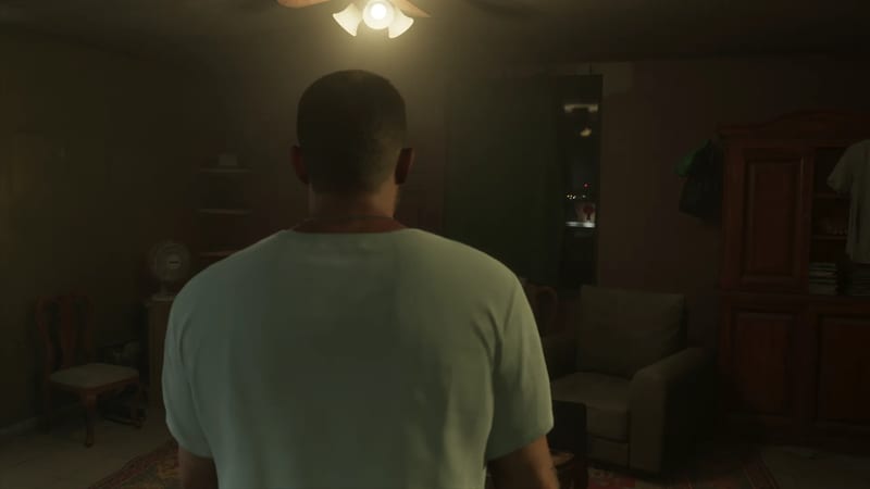 A man in a pale green T-shirt stands with his back to us in a dimly lit living room under a glowing ceiling light.
