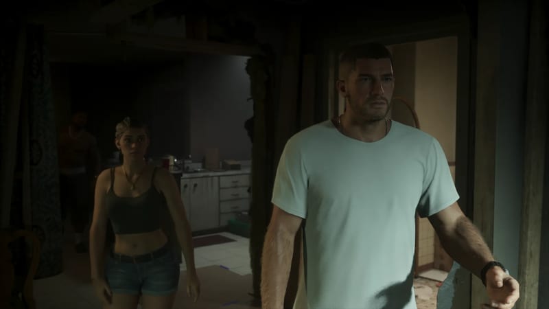 Jason Duval in a pale green t-shirt walks through a dim, run-down interior, with Lucia Caminos behind him and a kitchen beyond.
